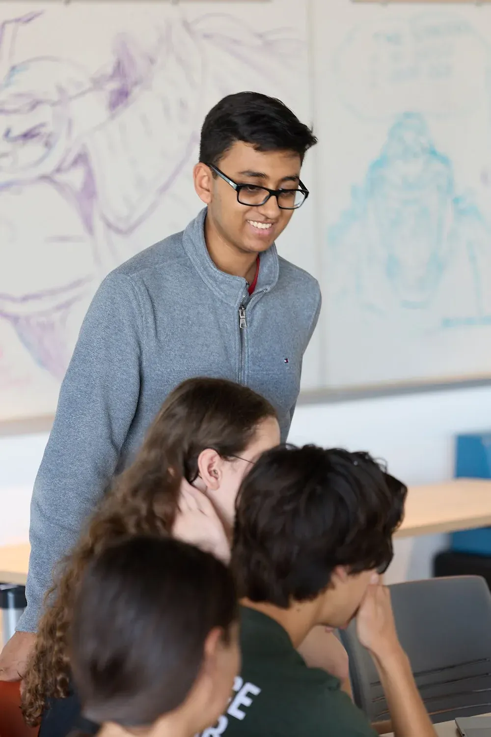 A smiling student with glasses stands among classmates working on laptops, with a whiteboard behind them